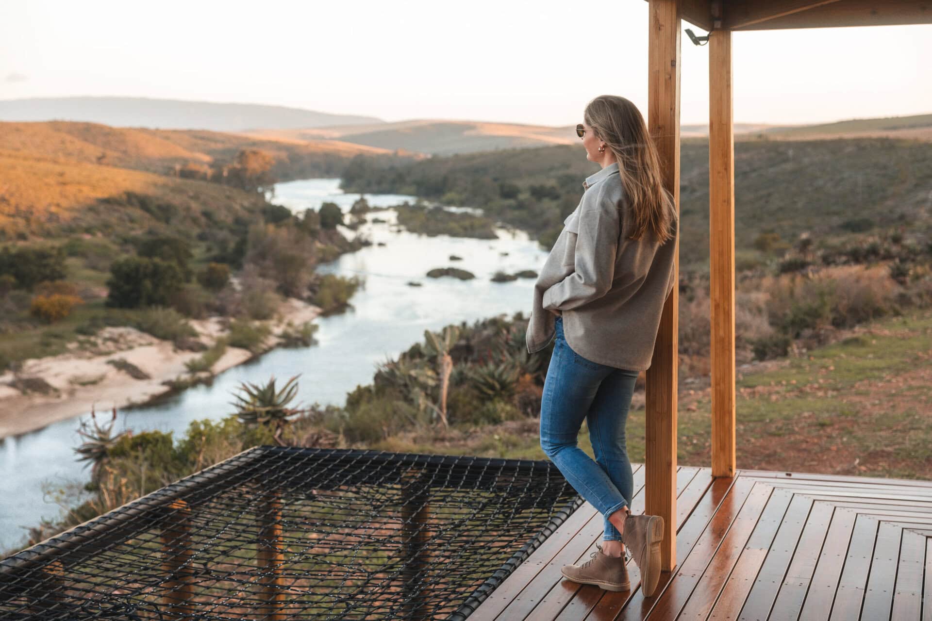 A woman stands on a wooden deck, gazing out over a peaceful river and rolling hills in the distance.